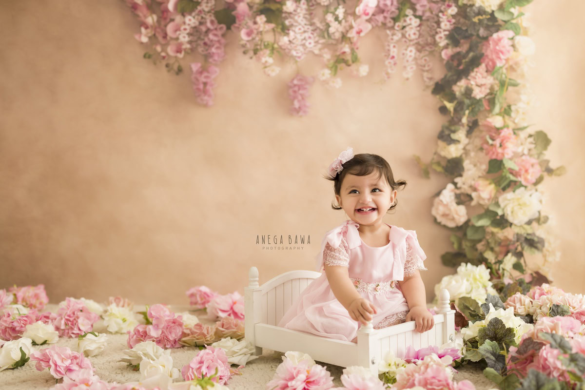 Capture the sweet innocence of your little princess with Anega Bawa's sitter photography in Delhi & Gurgaon. Nestled in a white cot against a soft peach backdrop, she wears a charming floral hair accessory, accentuating her adorable features. This delightful scene, adorned with floral fringes, perfectly encapsulates the innocence and beauty of your 10-month to 1-year-old daughter, creating cherished memories to last a lifetime.