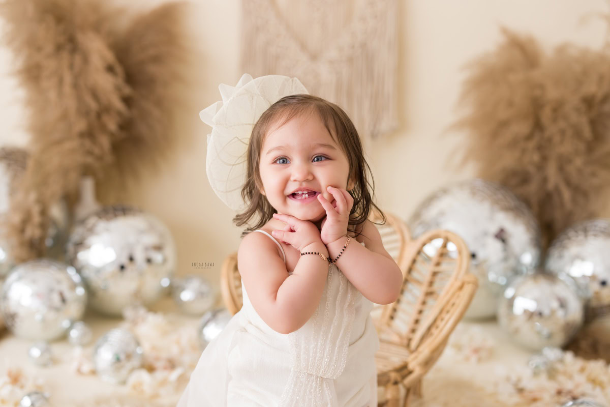Captured in this endearing toddler photoshoot by Anega Bawa in Delhi, Gurgaon, and Noida, a charming 10-month-old girl shines in a white dress paired with a cute hat. The scene is adorned with delicate discoballs and lush green bushes against a soothing beige backdrop. This picturesque setup exudes innocence and joy, beautifully encapsulating the playful spirit of childhood.