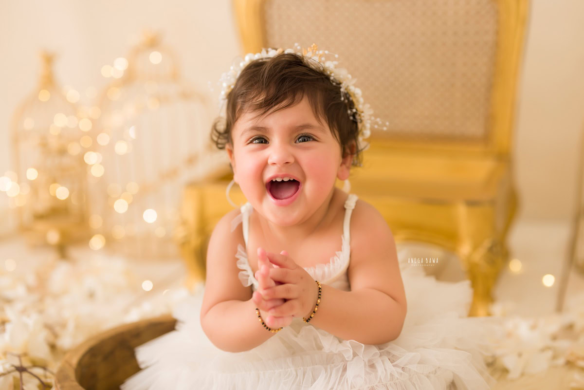 For a baby pre-birthday photoshoot in Delhi, Anega Bawa, Gurgaon, and Noida, capture the enchanting sight of a 10-month-old girl adorned in a white dress, seated on a golden chair amidst golden castles and fairy lights.