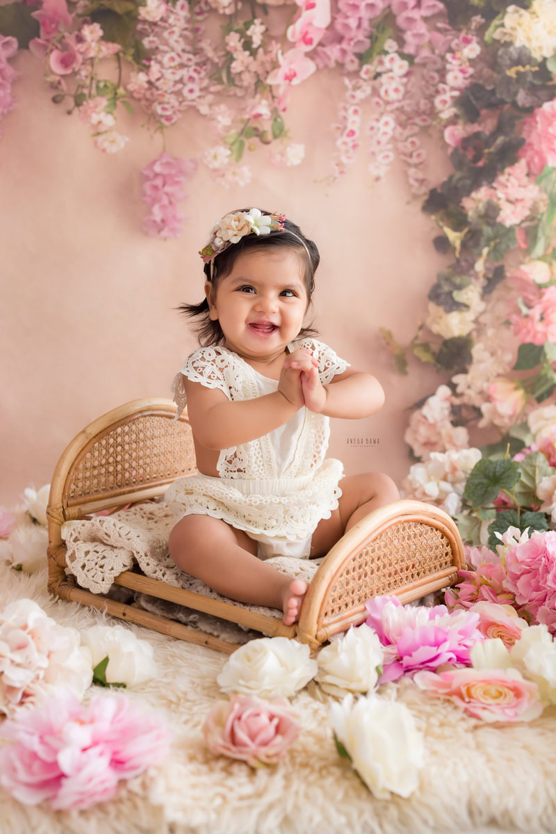 Immerse in the timeless charm of your little angel with Anega Bawa's sitter photography in Delhi & Gurgaon. Picture her nestled in a wooden cot against a delicate peach backdrop, surrounded by enchanting floral fringes. This serene setting beautifully captures the innocence and sweetness of your 10-month to 1-year-old daughter, creating cherished memories to treasure forever.