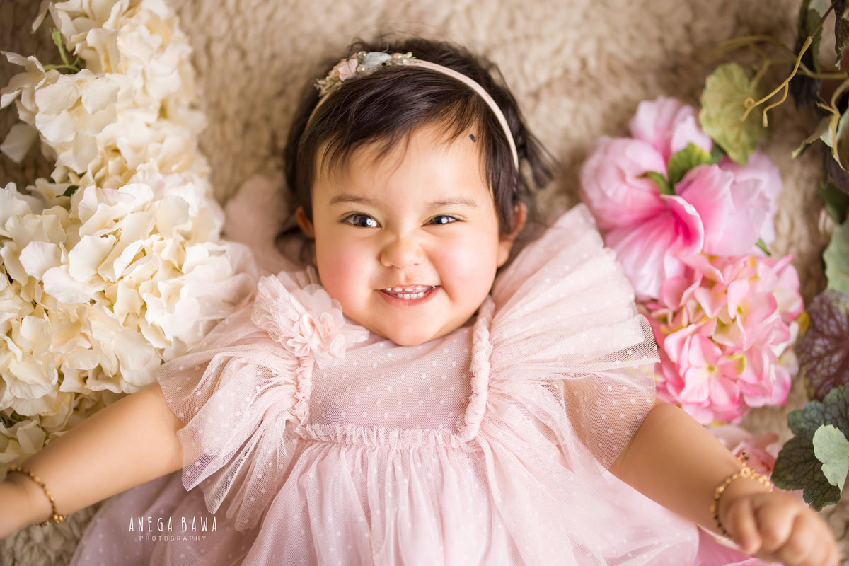 During a sitter photo shoot in Delhi, Gurgaon by Anega Bawa, a 10-month-old girl lays gracefully on a beige rug adorned with charming pink and white flowers. Dressed in an adorable pink dress and a cute headband, she exudes innocence and charm, creating a heartwarming scene.