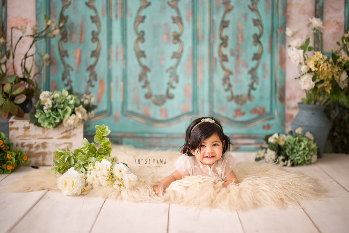In this captivating image captured by Anega Bawa, a beautiful 10-month-old girl lies on a cozy beige rug against a backdrop of a serene blue door. Surrounding her are delicate white flowers in a pot adorned with vibrant green leaves, adding a touch of natural elegance to the scene. Through her expert lens, the photographer has beautifully portrayed the innocence and serenity of childhood, creating a timeless moment to be cherished by the family. This enchanting photograph is a testament to the photographer's skill and creativity, offering a precious memory for years to come.