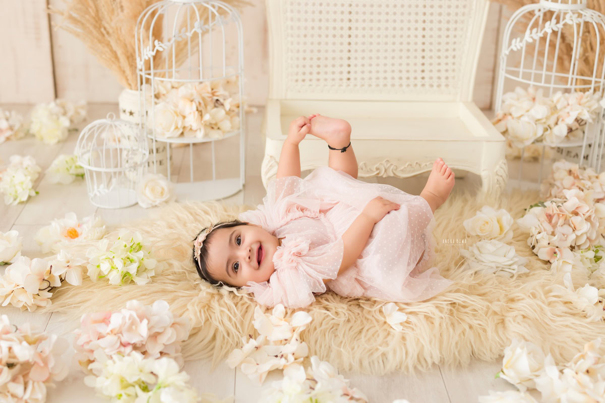 Indulge in the serene beauty of your little princess with Anega Bawa's sitter photography in Delhi & Gurgaon. Watch her lie gracefully on a beige rug amidst white castles, with a white wooden chair adding to the charm. Delicate flowers scattered on the floor complete the enchanting scene, capturing the innocence and elegance of your 10-month to 1-year-old daughter in timeless photographs to cherish forever.