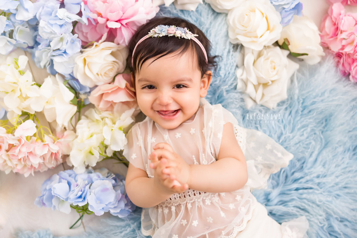 In a delightful sitter photo shoot in Delhi, Gurgaon by Anega Bawa, a 10-month-old girl lies on a soft blue rug, surrounded by vibrant multicolored flowers. With a lovely headband adorning her head, she adds a touch of innocence and sweetness to the scene.