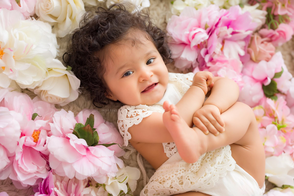 Girl lying on a bed of pink and white roses, striking a cute pose against a beautiful floral backdrop, marking her journey from 10 months to 1 year old during her first birthday photoshoot in Delhi by Anega Bawa, Gurgaon, Noida.