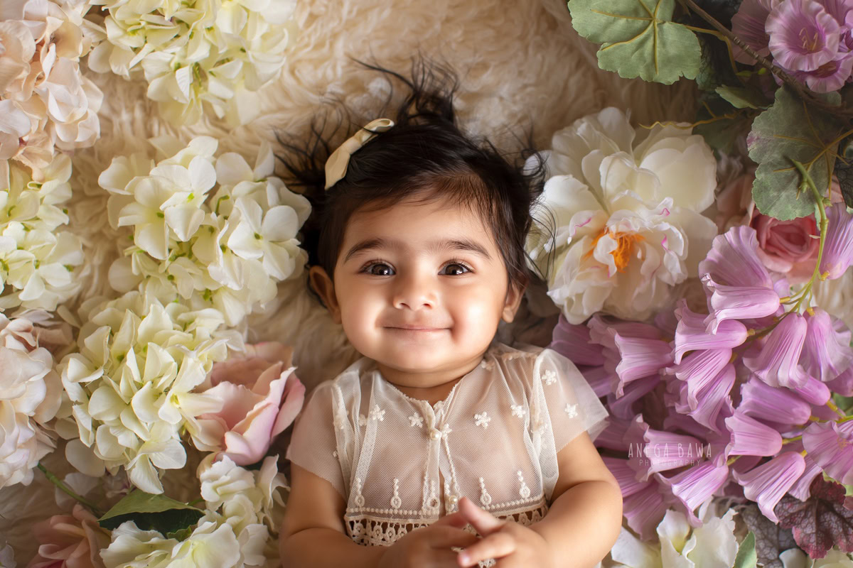 Captured during a heartwarming sitter photo session in Delhi, Gurgaon by Anega Bawa, this adorable 10-month-old girl strikes a charming pose amidst a backdrop adorned with vibrant multicolored flowers. Her innocence and joy radiate through the image, making it a cherished memory for years to come.