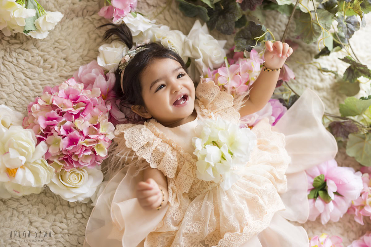 Girl lying on a beige rug surrounded by a bunch of flowers against a serene beige backdrop, marking her journey from 10 months to 1 year old during her first birthday photoshoot in Delhi by Anega Bawa, Gurgaon, Noida.