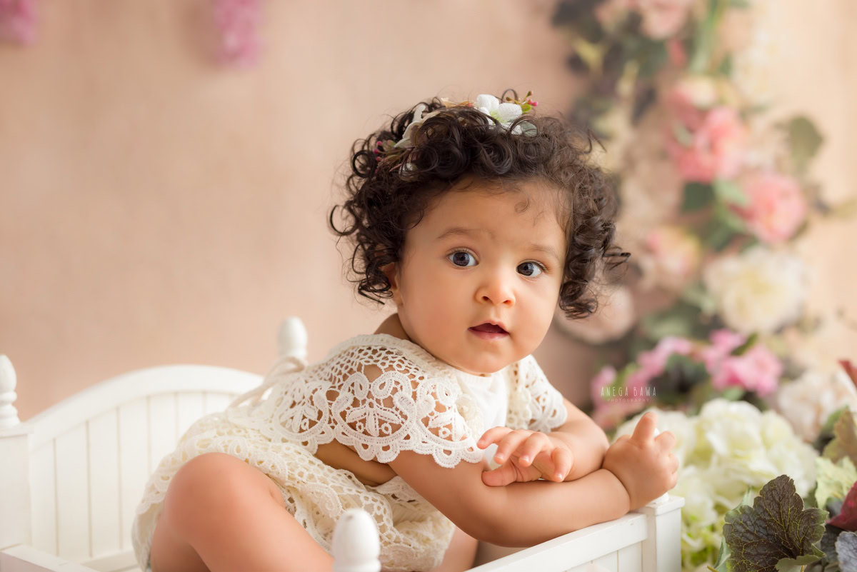 Capture the ethereal beauty of your little princess with Anega Bawa's sitter photography in Delhi & Gurgaon. Adorned in a delicate net dress, she sits gracefully on a white cot against a peach backdrop, surrounded by a charming floral fringe. These enchanting photographs, taken when your daughter is between 10 months and 1 year old, will be cherished memories for years to come.