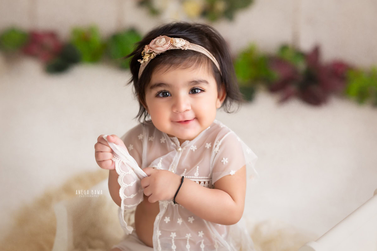 In this delightful photo by Anega Bawa, a charming 10-month-old girl sits on a beige rug, exuding innocence and joy. She wears a cute headband that accentuates her adorable features, while the leafy fringe on the floor adds a touch of natural beauty to the scene. Through the skillful lens of the photographer, this image captures the pure essence of childhood, offering a heartwarming glimpse into a moment of innocence and wonder. It's a precious memory to be cherished by the family for years to come.