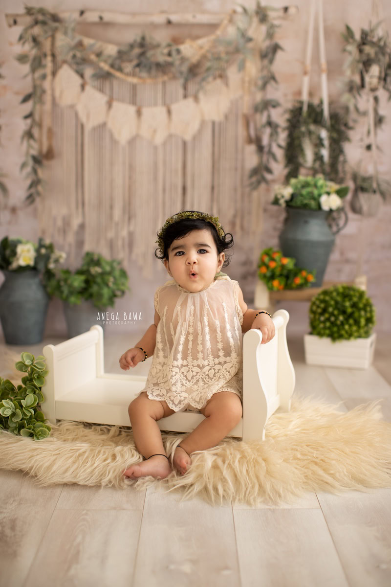 In this delightful sitter photo session in Delhi, Gurgaon by Anega Bawa, a charming 10-month-old girl is captured seated in a white cot, adorned with a net top. The scene is beautifully complemented by a beige rug and a blue vase alongside white pots against a serene beige backdrop.