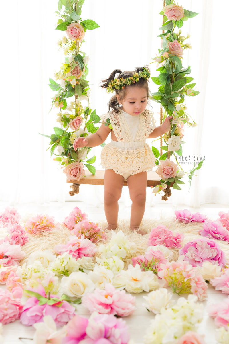 Witness the ethereal grace of your little princess with Anega Bawa's sitter photography in Delhi and Gurgaon. At 10 months to 1 year old, she radiates joy while seated on a leafy swing, surrounded by delicate flowers on the floor. Adorned with a tiara band and set against a pristine white backdrop, these enchanting images will capture the essence of her innocence and beauty for years to come.