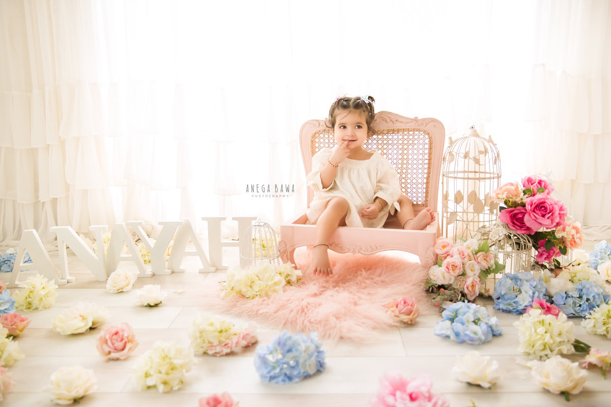 Delight in the precious moments with Anega Bawa Photography in Delhi and Gurgaon as your little princess graces a pink wooden chair amidst white castles and multicolored flowers. With a charming name frame adorning the floor, this captures the essence of her journey from 10 months to 1 year old.