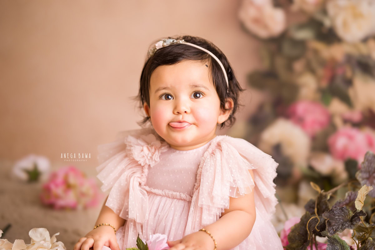 In this endearing sitter photo shoot by Anega Bawa in Delhi, Gurgaon, a lovely 10-month-old girl is seen wearing a pink dress against a soft peach backdrop. The scene is adorned with pink and white flowers, adding a touch of elegance to her adorable presence.