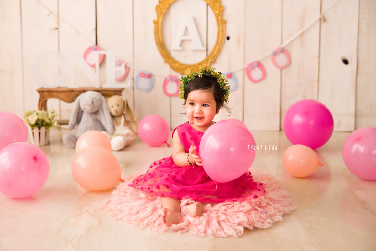 Captured by Anega Bawa, this delightful portrait features a 10-month-old girl in a pink dress, holding a teddy bear amidst peach and pink balloons. An alphabet frame on the wall adds educational charm.