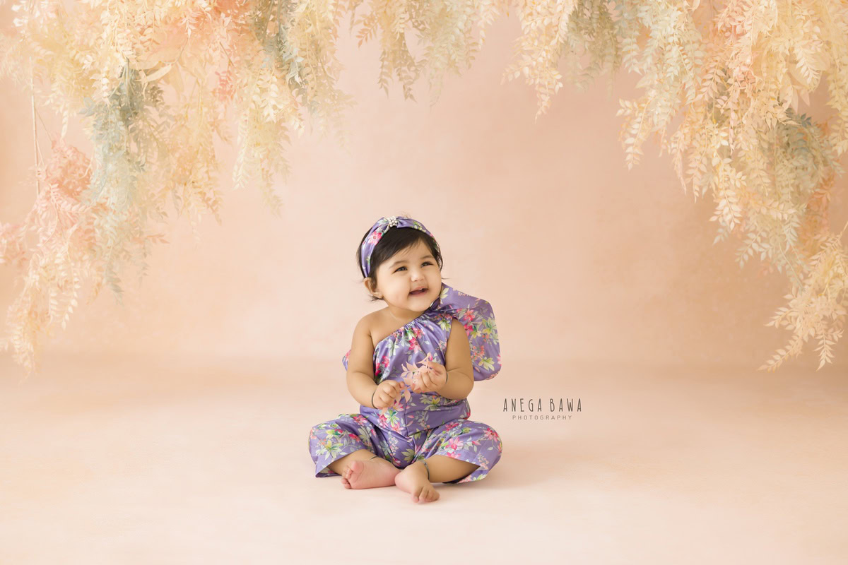 Indulge in the charm of your little angel with Anega Bawa's sitter photography in Delhi and Gurgaon. At 13 months, she sits gracefully, adorned in a one-shoulder dress and a delightful headband. Against a backdrop of lush peach foliage, her innocence shines through, creating timeless memories you'll cherish forever.