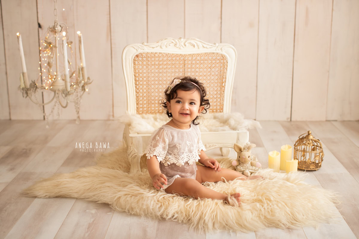 At 10 months old, this seated girl, photographed by Anega Bawa, basks in the warm glow of candlelight while perched on a beige rug against a serene beige backdrop.