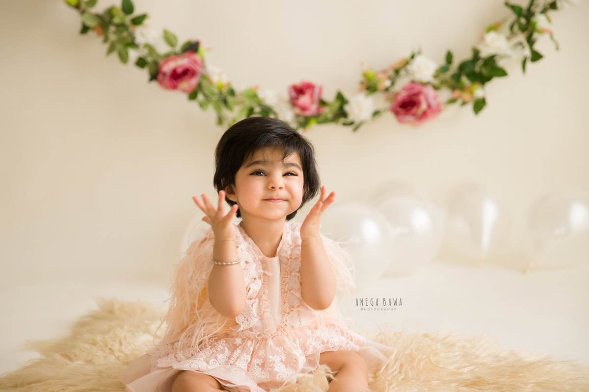 Celebrate your little girl's journey from 10 months to 1 year old with Anega Bawa Photography in Delhi and Gurgaon. Watch her adorable charm unfold as she sits on a beige rug surrounded by floral fringe and balloons against a serene beige backdrop.
