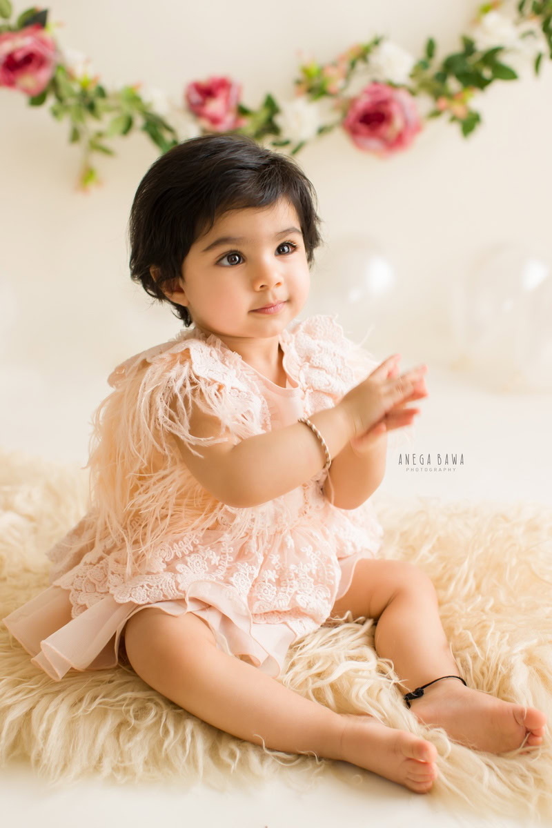 Capture the precious moments of your growing princess with Anega Bawa Photography in Delhi and Gurgaon. Adorned in a floral fringe setting, she sits on a beige rug against a soft beige backdrop, showcasing her adorable charm at 10 months to 1 year old.