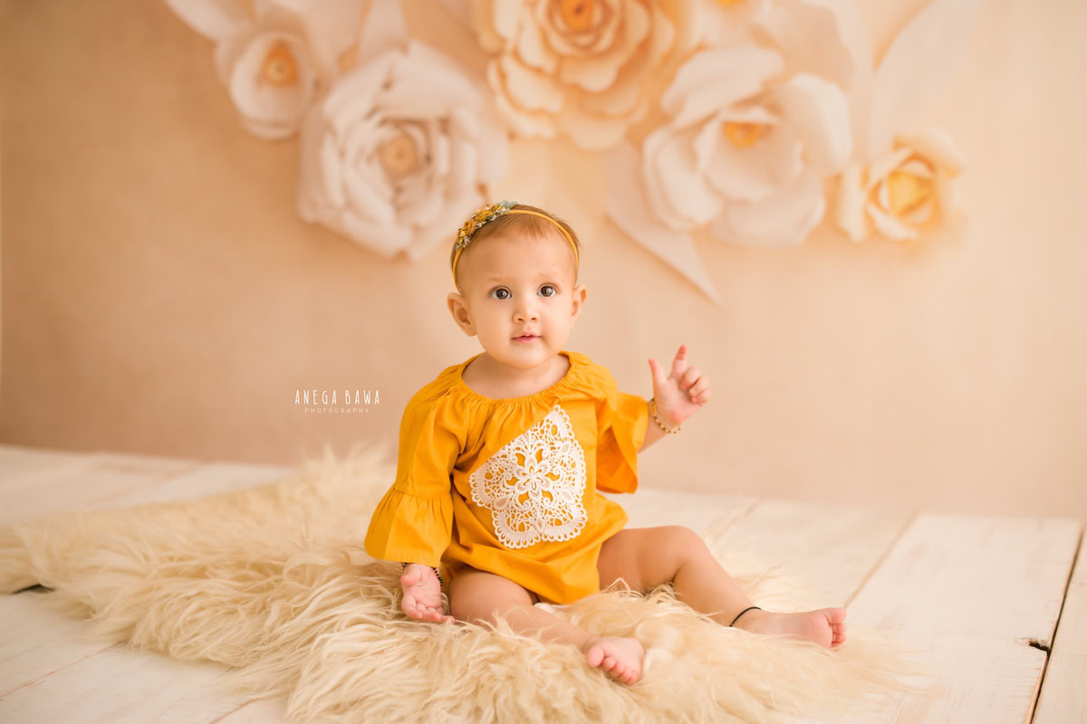 Create timeless memories of your little princess with Anega Bawa Photography in Delhi and Gurgaon. Adorned in a charming floral setting, she sits gracefully on a beige rug against a soft peach backdrop, radiating her adorable charm at 10 months to 1 year old.