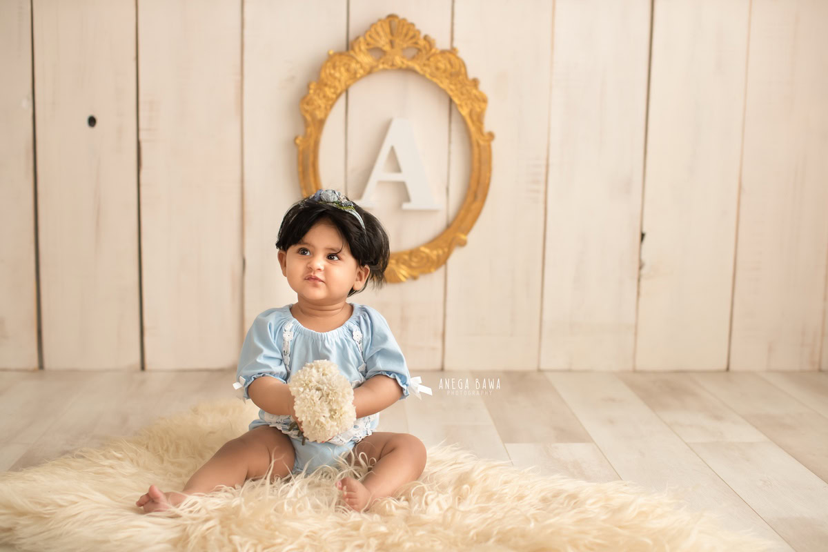 For a captivating baby pre-birthday photoshoot in Delhi, Anega Bawa, Gurgaon, and Noida, picture a 10-month-old girl seated gracefully on a beige rug against a backdrop of golden frame on the wall, creating a mesmerizing ambiance.