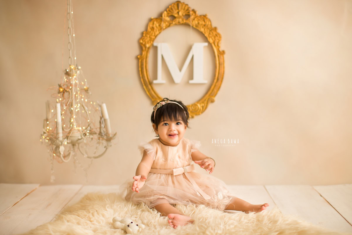 For an enchanting baby pre-birthday photoshoot in Delhi, Anega Bawa, Gurgaon, and Noida, envision a 10-month-old girl seated on a beige rug, adorned by the glow of golden lights from a candle stand, with a golden name frame adorning the wall in the background, casting a warm and magical aura.