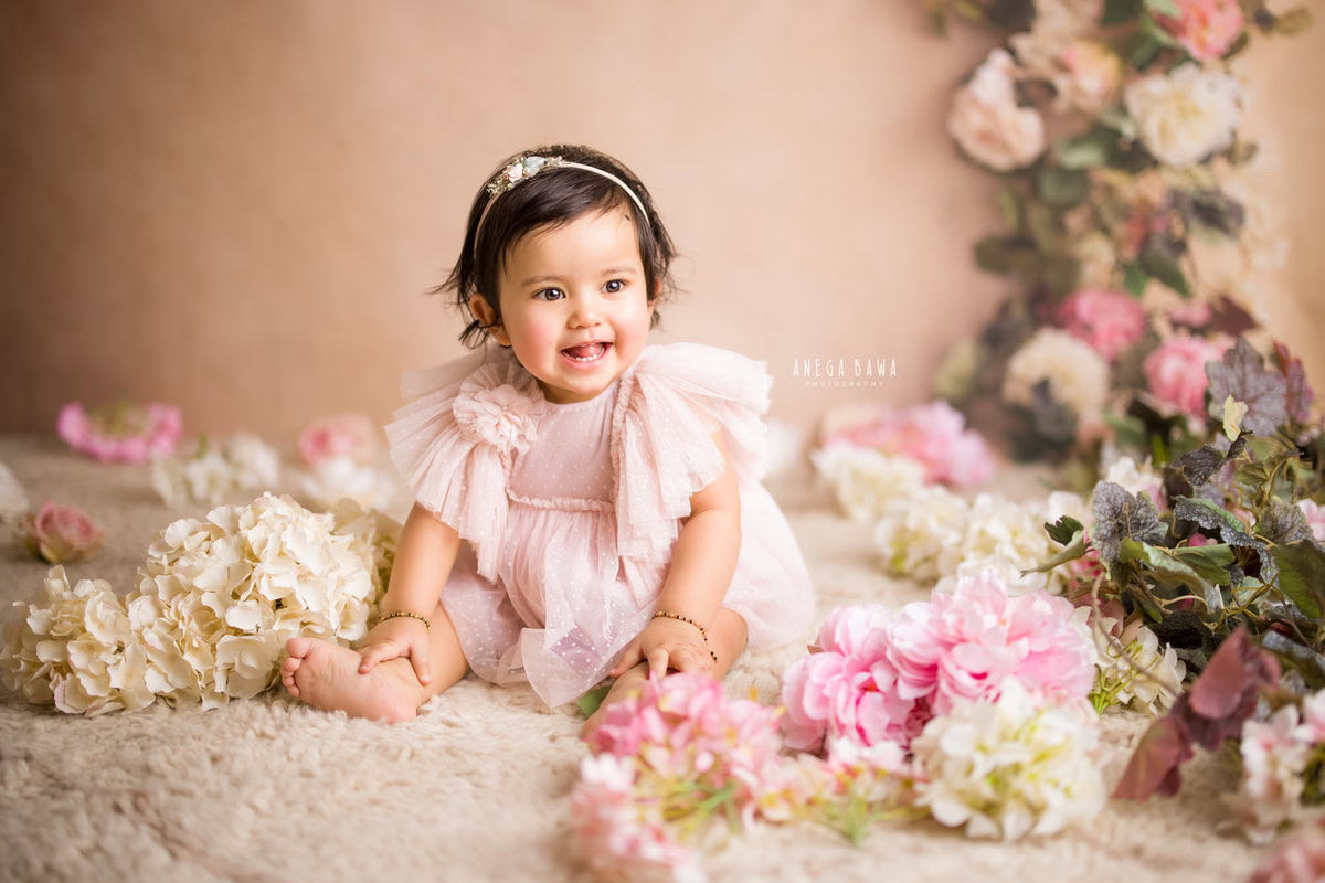 Captured during a sitter photo shoot in Delhi, Gurgaon by Anega Bawa, this charming 10-month-old girl is seated on a beige rug against a peach backdrop. Surrounding her are multicolored flowers, creating a delightful and cheerful atmosphere for the shoot.