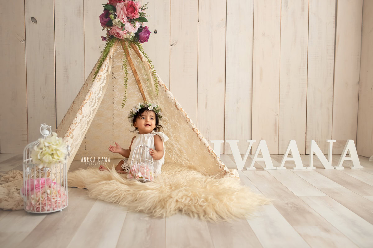 Create timeless memories with Anega Bawa Photography in Delhi and Gurgaon as your little princess sits elegantly on a beige rug amidst a net tent and white castle props. Against a soft beige backdrop adorned with floral bunches, her innocence shines through in every frame at 10 months to 1 year old.