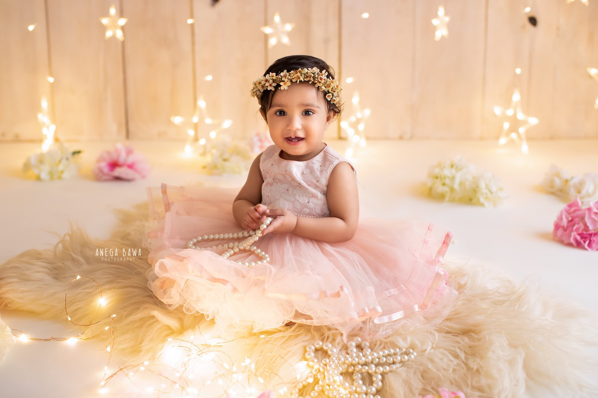 Captured by the talented Anega Bawa in Delhi-Gurgaon, this delightful image features a 9-month-old girl seated on a beige rug, adorned with star-shaped fairy lights and scattered flowers. The serene beige backdrop adds to the enchanting ambiance of the scene.