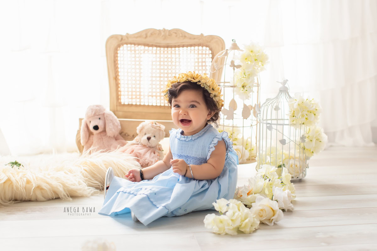 For a delightful baby pre-birthday photoshoot in Delhi, Anega Bawa, Gurgaon, and Noida, picture a 9-month-old girl seated on a beige rug surrounded by cuddly teddy bears and whimsical white castles. White flowers accentuate the scene against a serene white backdrop, creating a charming and picturesque setting.