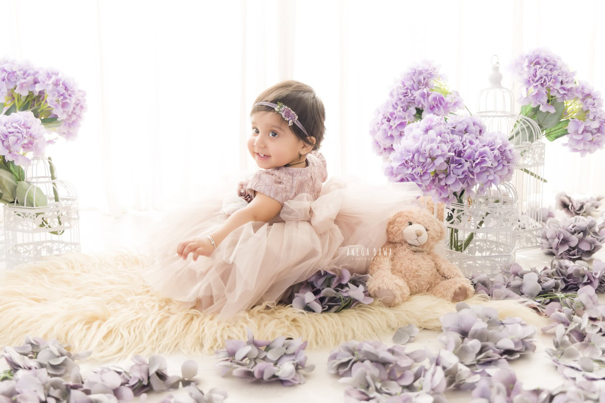 Girl seated on a beige rug amidst white castles and lavender flowers against a pristine white backdrop, marking her journey from 9 months to 1 year old during her first birthday photoshoot in Delhi by Anega Bawa, Gurgaon, Noida.