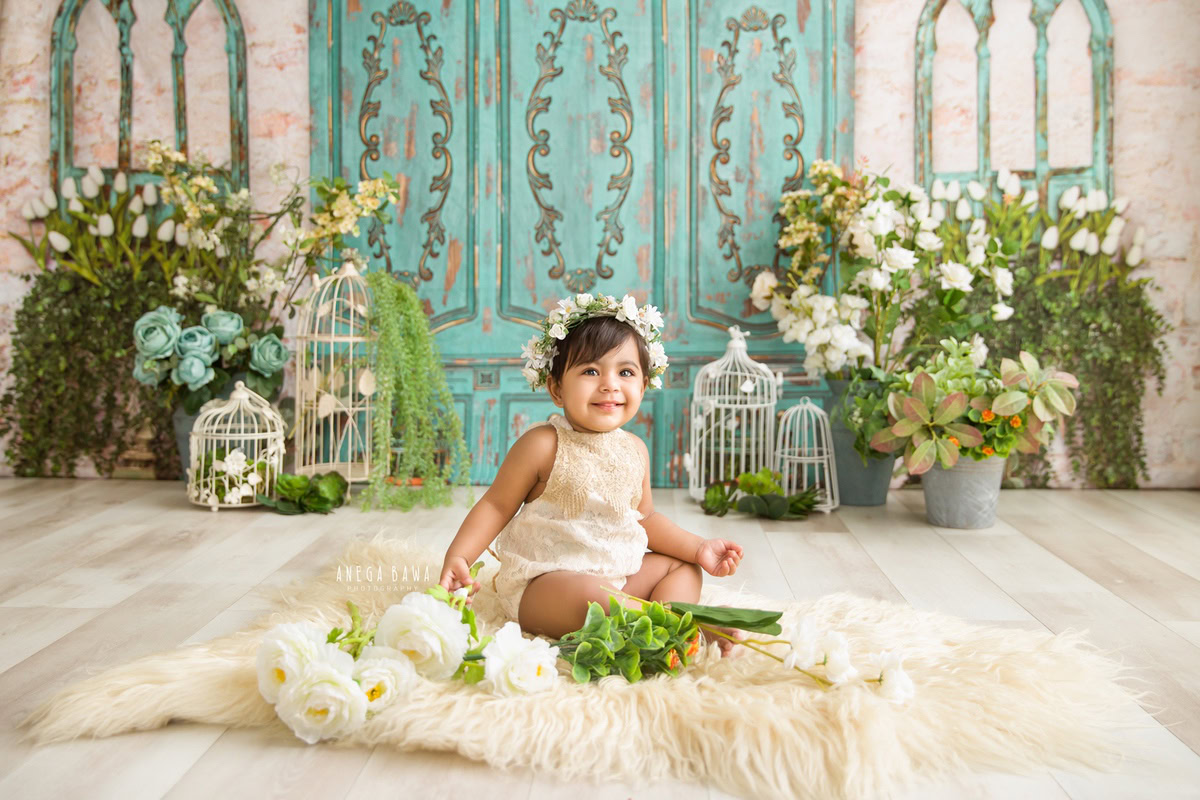 Capture the pure innocence of your little princess with Anega Bawa Photography in Delhi and Gurgaon. Seated gracefully on a beige rug surrounded by white castles and delicate flowers, against a lush green and beige backdrop, she creates enchanting memories at 9 months to 1 year old.