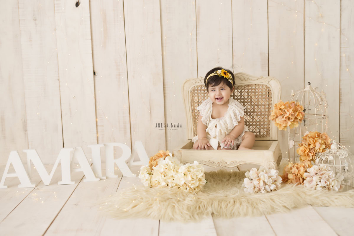 Capture the enchantment of a baby pre-birthday photoshoot in Delhi, Anega Bawa, Gurgaon, and Noida with a 9-month-old girl seated gracefully on a beige wooden chair. The beige backdrop sets the stage for a picturesque scene adorned with a name frame resting on the floor. Delicate flowers and charming castles embellish the beige rug, adding to the delightful ambiance of this captivating moment.