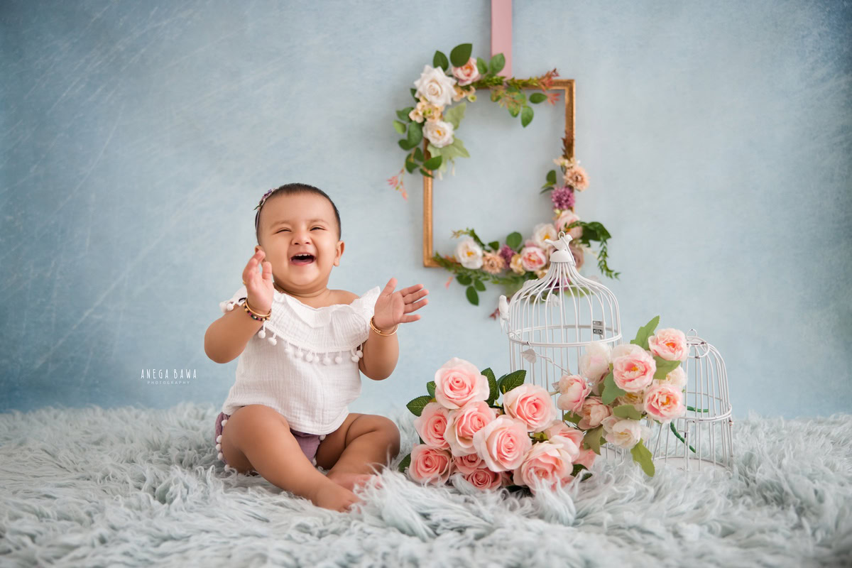 In this adorable capture from a sitter photo shoot in Delhi, Gurgaon by Anega Bawa, a sweet 9-month-old girl is seated on a blue rug against a blue backdrop. A floral wooden frame on the wall adds a charming touch, while white castles and peach flowers complete the scene, adding a whimsical flair to the setup.