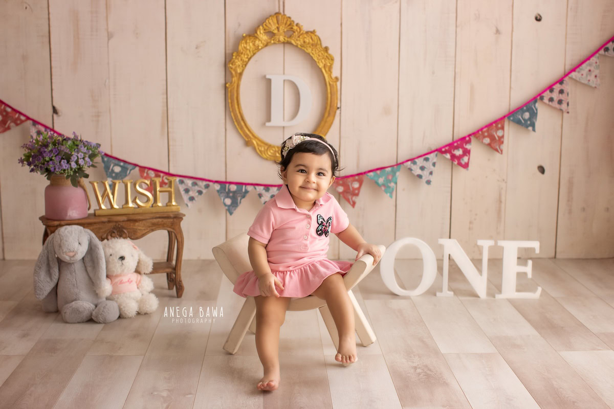 In a charming capture by Anega Bawa in Delhi-Gurgaon, a 9-month-old girl is seated on a curvy chair, surrounded by colorful teddy bears and a vibrant alphabet frame adorning the wall. This delightful scene is enhanced by the playful addition of a colorful fringe, adding to the joyous atmosphere of the moment.