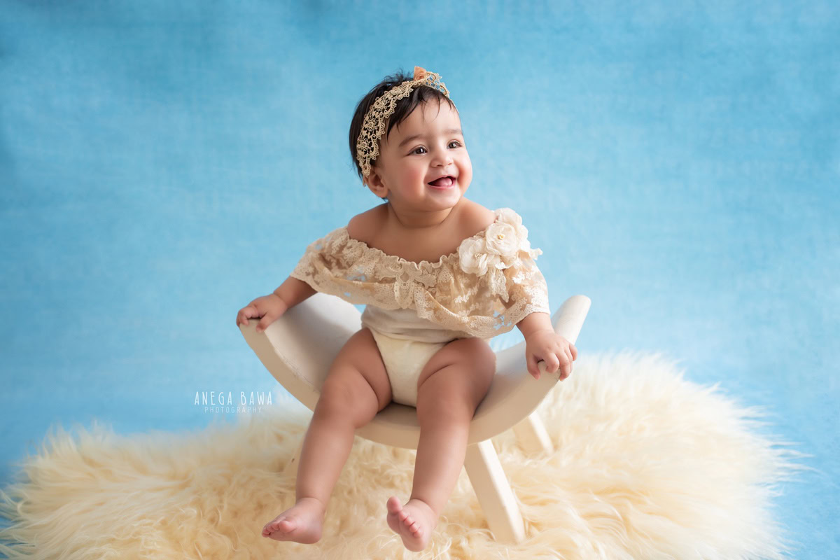 Celebrate the precious moments of your little princess's journey with Anega Bawa Photography in Delhi and Gurgaon. Adorably seated on a curvy chair atop a beige rug, against a serene blue backdrop, she shines bright at 9 months to 1 year old.