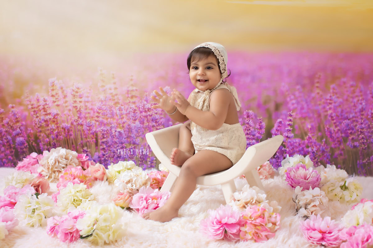 In a picturesque moment captured by Anega Bawa in Delhi-Gurgaon, a 9-month-old girl sits gracefully on a curvy chair against a soothing lavender backdrop. Surrounding her are delicate pink and white flowers, adding a touch of elegance to the scene. This enchanting composition beautifully captures the innocence and charm of the little one's early years.