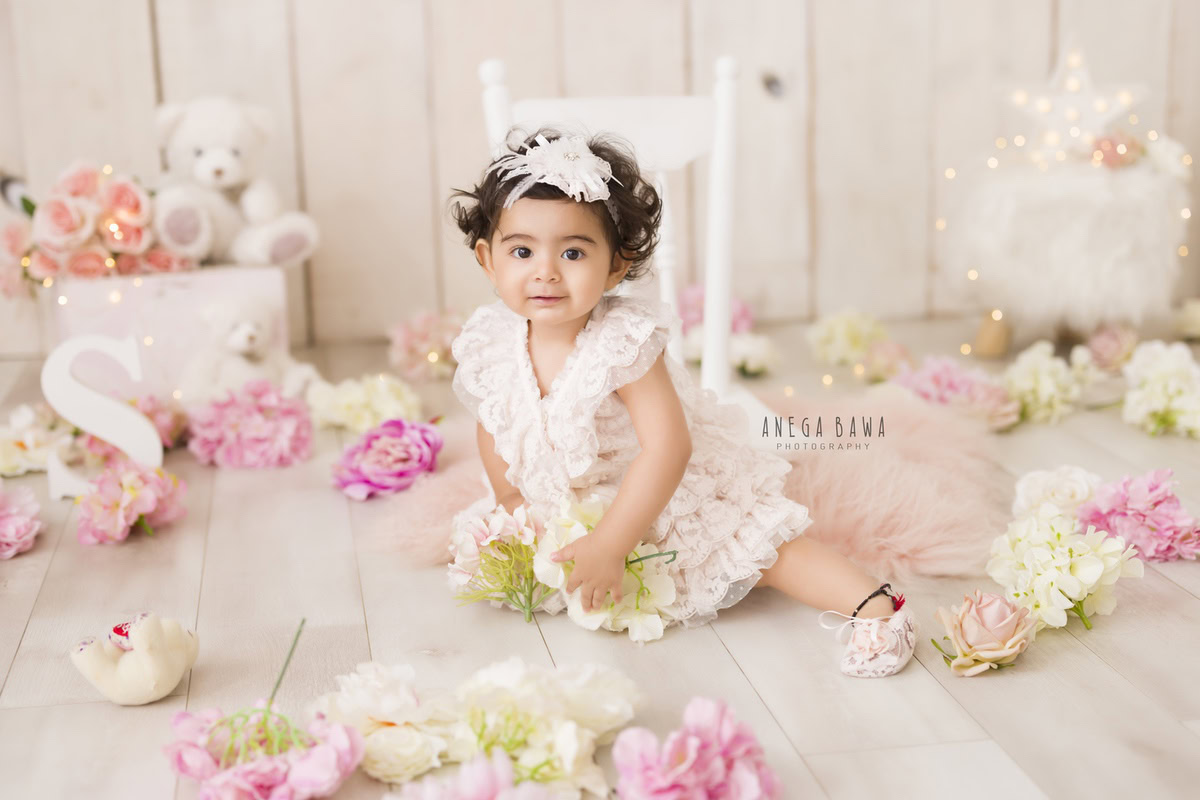 Girl seated on a white chair adorned with cute booties, surrounded by multicolored flowers and a teddy bear, with fairy lights illuminating the scene, marking her journey from 9 months to 1 year old during her first birthday photoshoot in Delhi by Anega Bawa, Gurgaon, Noida.