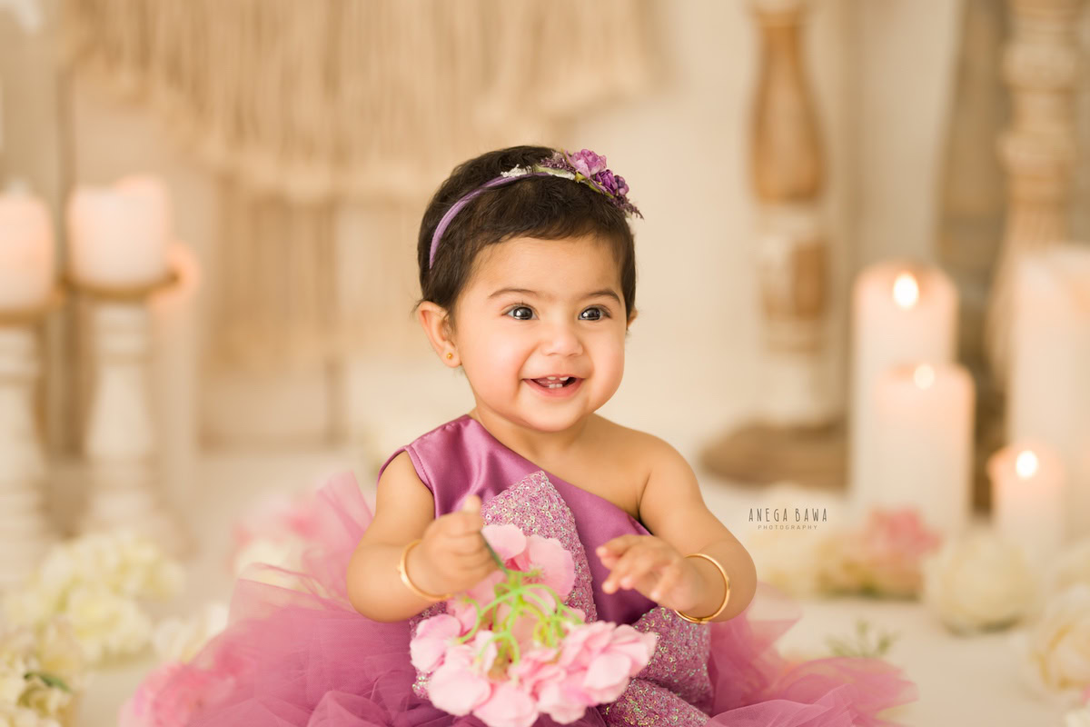 In this delightful moment captured during a sitter photo shoot in Delhi, Gurgaon by Anega Bawa, a 9-month-old girl sits gracefully against a beige backdrop. Holding a flower in her hand, she adds a touch of innocence and charm to the scene. Surrounding her are candle stands and scattered white flowers on the floor, creating a serene and picturesque setting.
