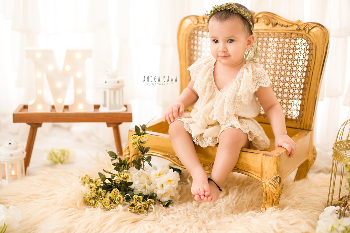 Witness the enchanting beauty of your little princess in a captivating photoshoot by Anega Bawa Photography in Delhi and Gurgaon. Seated gracefully on a golden chair amidst a bed of beige rugs and golden-white flowers, against a pristine white backdrop, she radiates joy and innocence at 9 months to 1 year old.