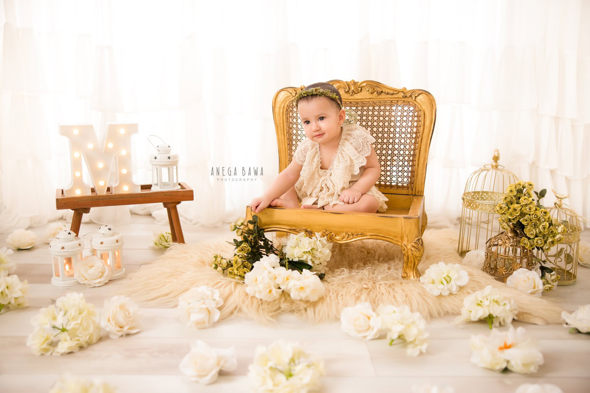 Capture the pure essence of your little princess with Anega Bawa Photography in Delhi and Gurgaon. Adorned with a golden tiara band and surrounded by white flowers and golden castles, she sits elegantly against a serene white backdrop, creating timeless memories at 9 months to 1 year old.