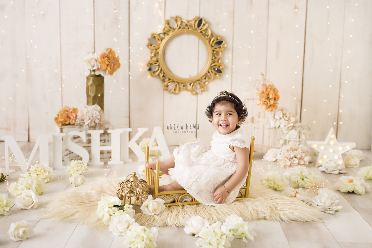 In a delightful scene framed by Anega Bawa in Delhi-Gurgaon, a 9-month-old girl sits snugly on a golden cot against a warm beige backdrop. Adorning the wall behind her is a charming frame, while vibrant white and orange flowers add a pop of color to the serene setting. This captivating image perfectly encapsulates the innocence and sweetness of the little one's early years.