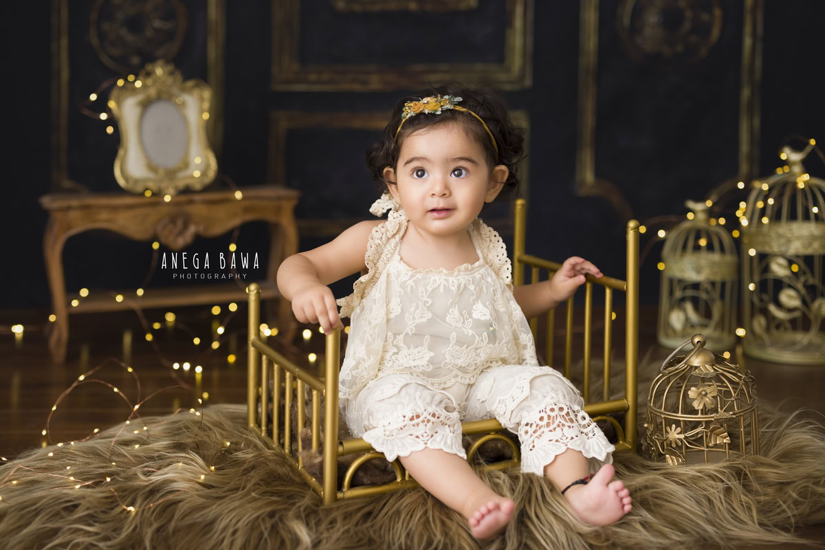 In a picturesque moment captured by Anega Bawa in Delhi-Gurgaon, a 9-month-old girl is seated gracefully on a golden cot, surrounded by the soft warmth of fairy lights against a rich brown backdrop. Beneath her feet lies a plush brown rug, while golden castles add a touch of whimsy to the enchanting scene. This beautifully composed photograph captures the essence of childhood innocence and wonder, making it a timeless keepsake for years to come.
