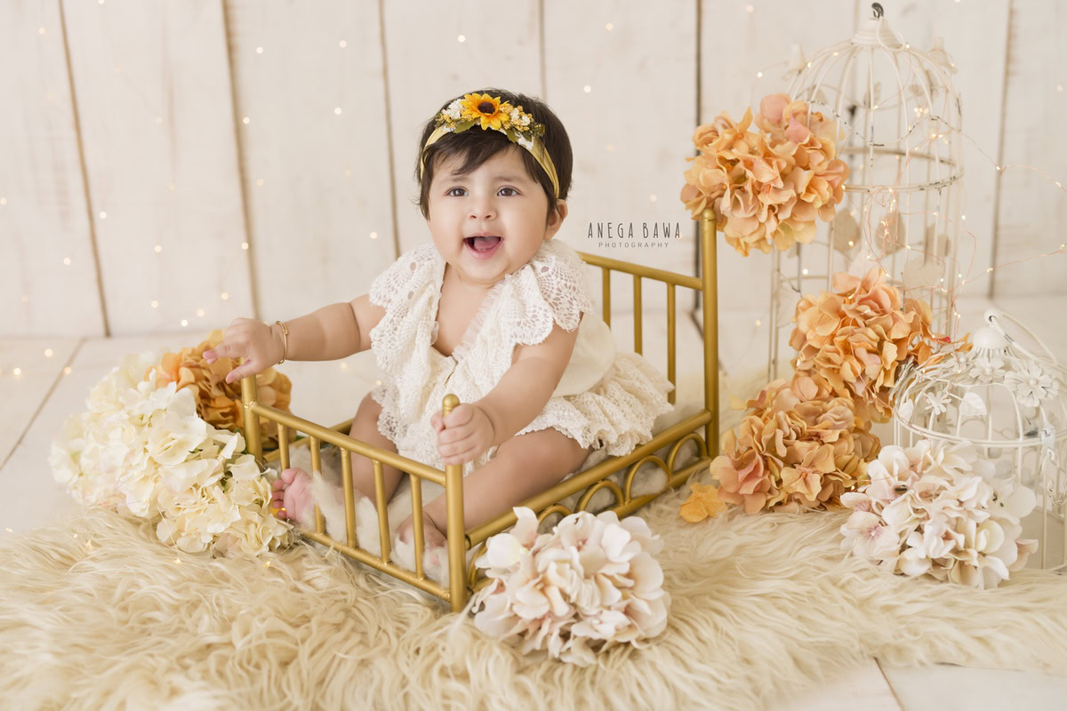 Immerse yourself in the magic of a baby pre-birthday photoshoot in Delhi, Anega Bawa, Gurgaon, and Noida as a 9-month-old girl graces a golden cot. Adorned in a delicate net dress and a charming sunflower headband, she sits amidst enchanting castles and vibrant orange flowers against a serene beige backdrop. Capture this precious moment in time, where innocence meets elegance, in a scene that radiates warmth and joy.