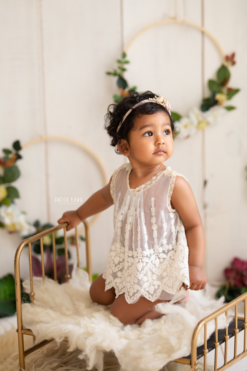 During a sitter photo shoot in Delhi, Gurgaon by Anega Bawa, a captivating moment was captured as a 9-month-old girl sits elegantly on a golden cot. The scene is adorned with a white rug and circular leafy frames on the wall, creating a serene and enchanting ambiance against a pristine white backdrop. This delightful composition beautifully encapsulates the innocence and grace of infancy.