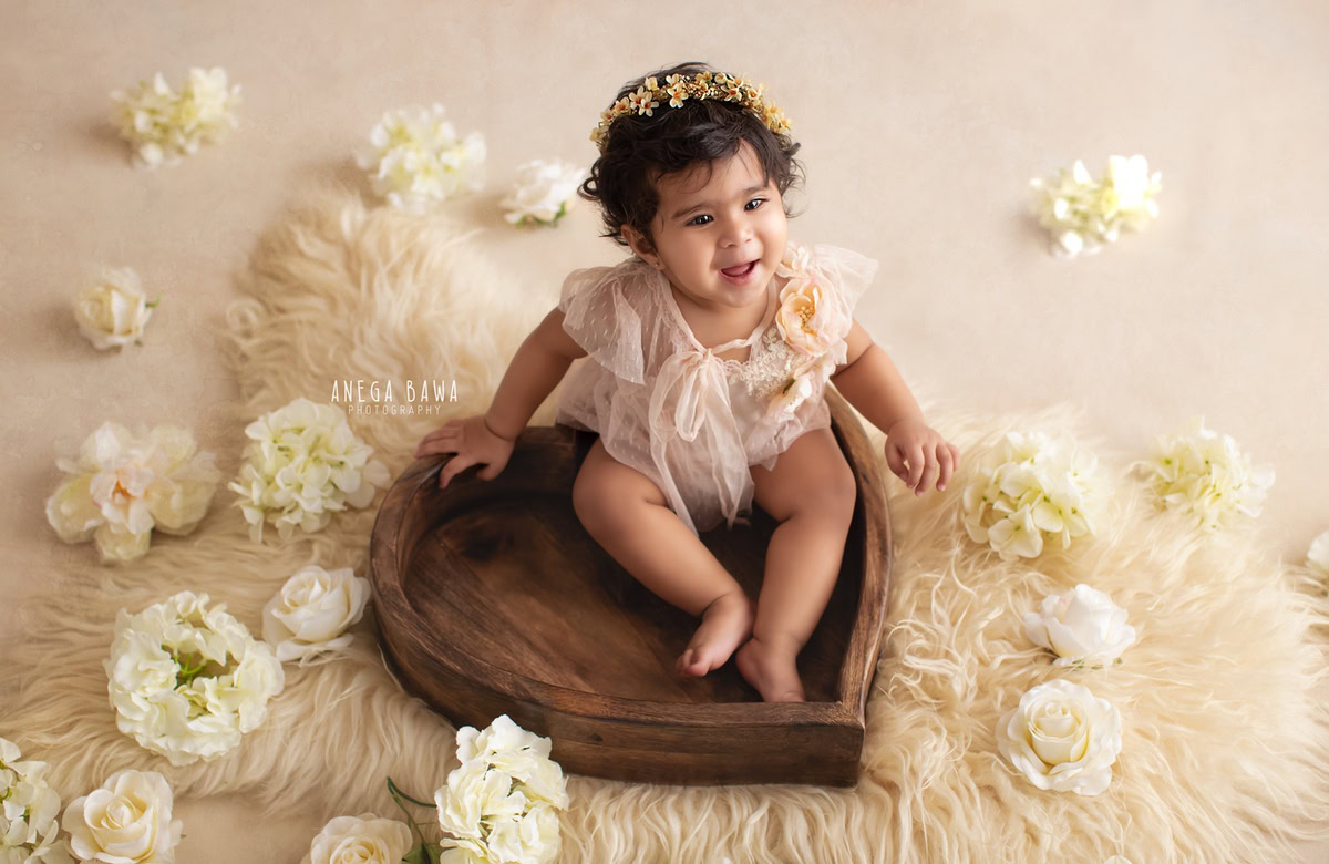 In a charming sitter photo session in Delhi, Gurgaon by Anega Bawa, a heartwarming moment unfolds as a 9-month-old girl sits gracefully in a heart-shaped wooden basket. The scene is adorned with a beige rug and delicate white flowers, set against a pristine white backdrop. This endearing setup captures the innocence and beauty of infancy, creating a timeless memory to cherish.