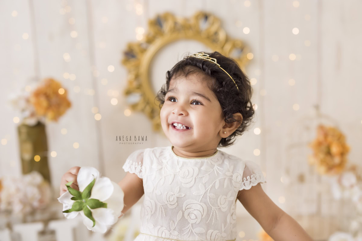 In a captivating composition by Anega Bawa in Delhi-Gurgaon, a 9-month-old girl sits serenely against a soothing beige backdrop, her delicate fingers holding a vibrant flower. The scene is bathed in the soft glow of fairy lights, casting a magical ambiance around her. A delicate flower vase adds an extra touch of elegance to the enchanting tableau. This evocative photograph beautifully captures the innocence and beauty of early childhood, making it a cherished memento for years to come.
