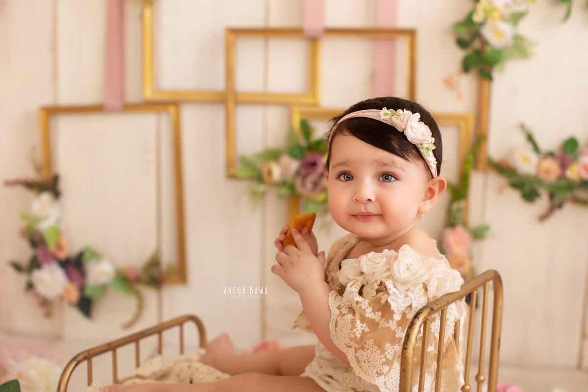 Celebrate the innocence of your little angel with Anega Bawa Photography in Delhi and Gurgaon. Nestled in a golden cot against a gentle beige backdrop adorned with floral frames, she creates captivating memories at 9 months to 1 year old.