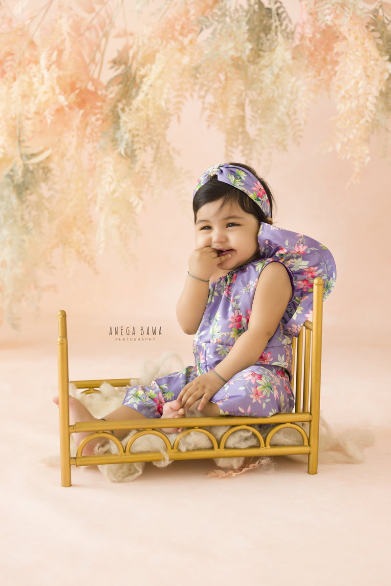 Girl seated in a golden cot against a backdrop adorned with leafy peachy hues, marking her journey from 9 months to 1 year old during her first birthday photoshoot in Delhi by Anega Bawa, Gurgaon, Noida.