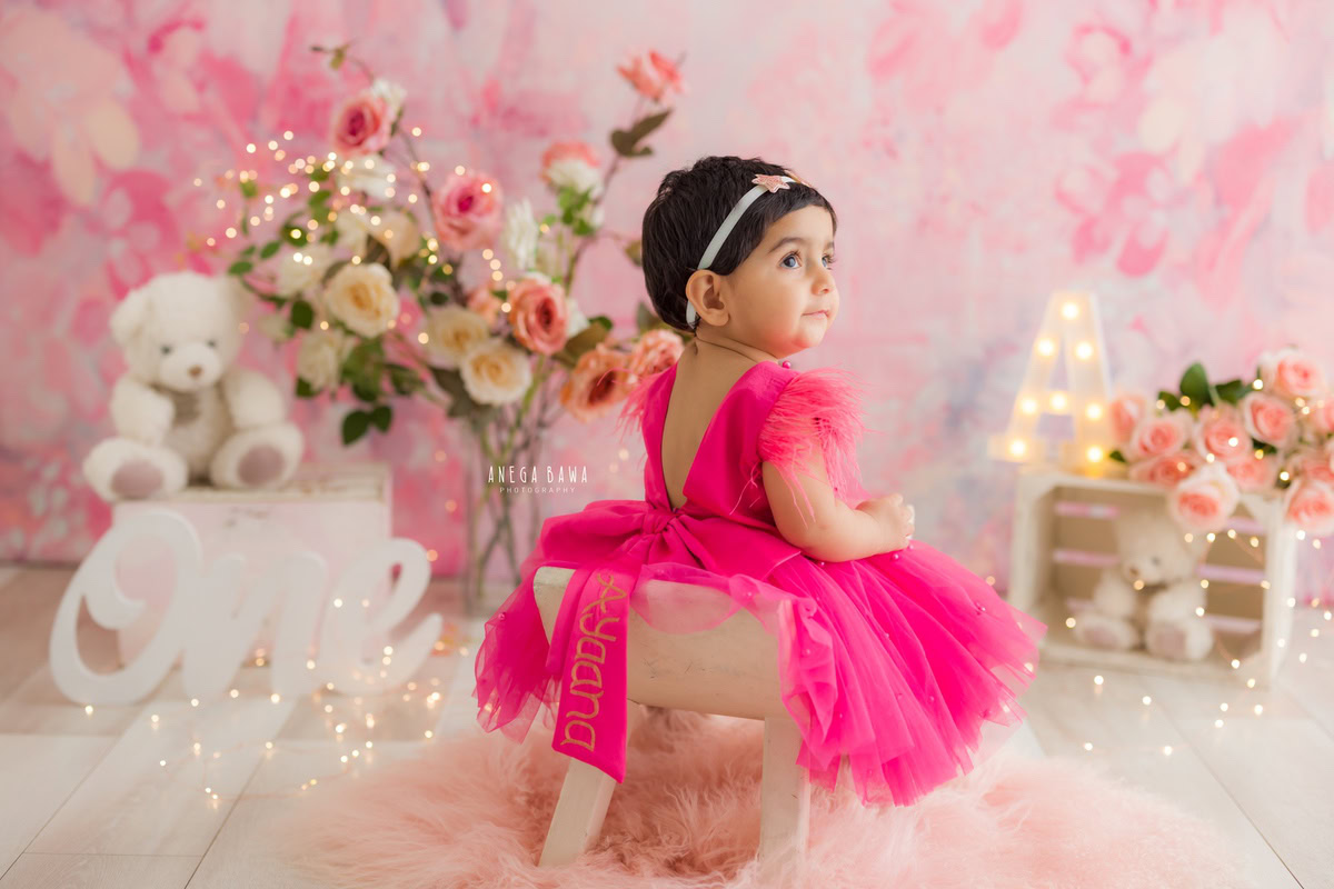 Girl seated in a tub adorned with a pink dress against a charming pink backdrop, surrounded by teddy bears and a bunch of flowers, with fairy lights adding a magical touch, marking her journey from 8 months to 1 year old during her first birthday photoshoot in Delhi by Anega Bawa, Gurgaon, Noida.