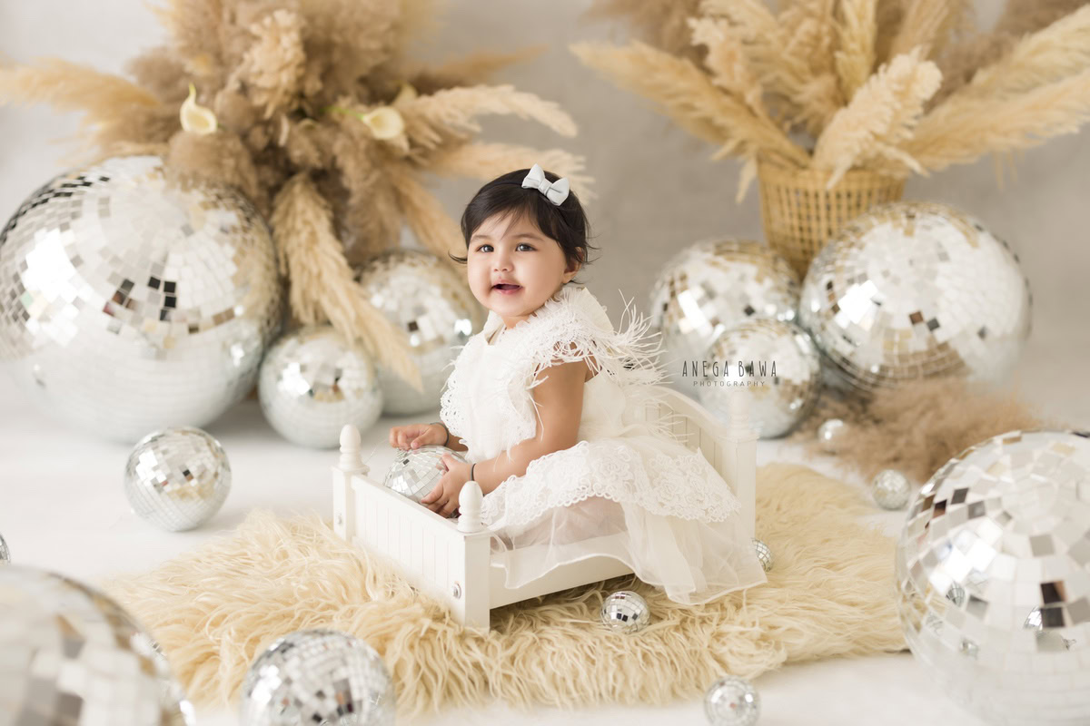 Delight in the radiance of your 8-month-old angel, seated serenely in a pristine white cot. With a beige rug beneath her and a backdrop of twinkling disco balls amidst lush bushes, each snapshot captures her innocence and charm. Experience the enchantment of sitter photography in Delhi and Gurgaon with Anega Bawa, where every moment is a treasure to behold.